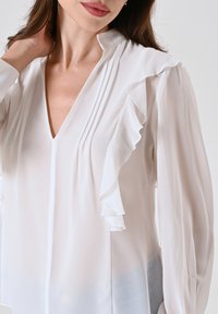 Blusa bianca realizzata in tessuto leggero, con scollatura a V, design plissettato e dettagli arricciati lungo la parte anteriore e le maniche.