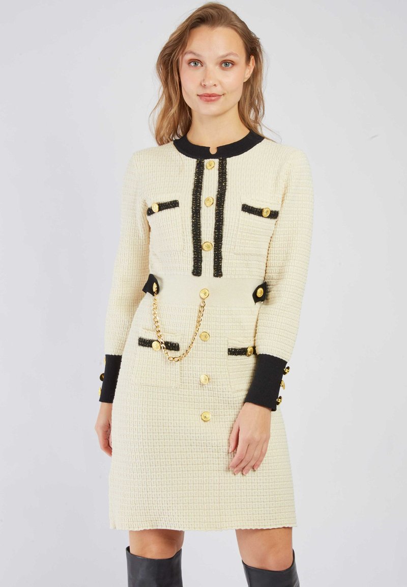 Kremowa sukienka o teksturze z czarnym wykończeniem, złotymi guzikami, dwiema przednimi kieszeniami oraz łańcuszkiem w talii. Długie rękawy z kontrastowymi mankietami.