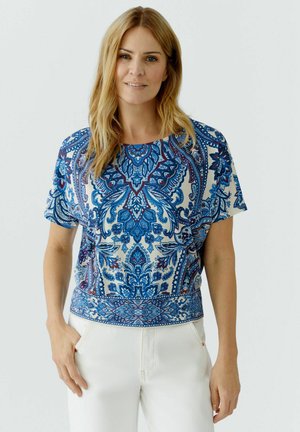 Woman wearing a short-sleeve blouse with blue paisley patterns and white pants, standing with one hand in pocket against plain background.