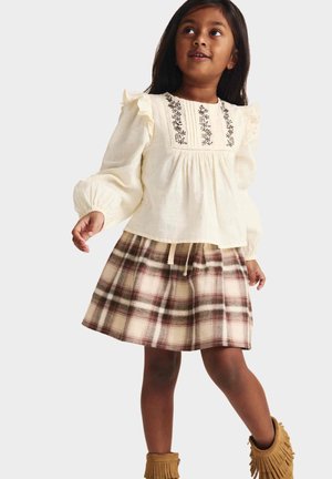 Ragazza giovane che indossa una camicetta color crema con ricami floreali e volant, gonna a quadri e stivaletti marroni con frange, in piedi e guarda verso l'alto.