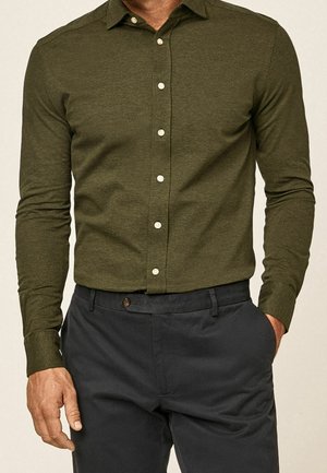 Mies pukeutuneena oliivinvihreään pitkähihainen nappipaitaan, joka on tungettu tummanharmaisiin housuihin, ja toisessa kädessä taskussa.