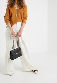 Kvinna som bär en orange tröja och vita vida byxor, håller en svart handväska med guldkedjor, står på en vit golv.