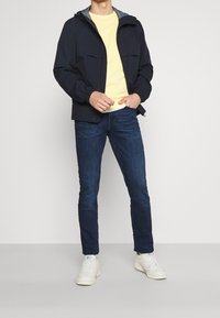 Mörkblå lättviktsjacka med huva och elastiska muddar, bärs över en gul t-shirt, tillsammans med mörka jeans och vita sneakers.