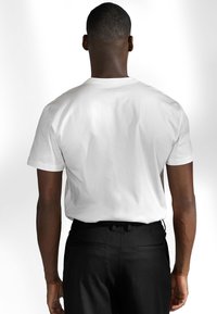 Vit bomull t-shirt med kort ärm, visad bakifrån, parat med svarta byxor. Ren design utan synliga grafiker eller logotyper.