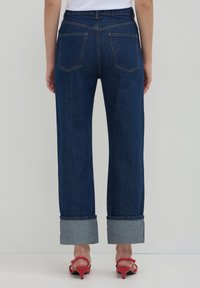 Jean en denim bleu foncé à la coupe décontractée, avec des manches retroussées et deux poches arrière. Porté avec des sandales à talons rouges.