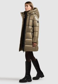 Manteau rembourré de longueur genou en vert olive métallique avec une capuche, présentant une fermeture éclair avant, des poches latérales et des accents noirs. Stylisé avec des bottes noires.