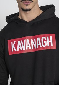 Čierna mikina s kapucňou vyrobená z hrubej tkaniny, ktorá má na sebe odvážny červený obdĺžnikový nášivok so bielym textom "KAVANAGH" v strede.