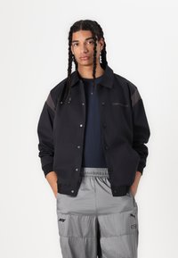 Chaqueta negra de botones con acentos grises en los hombros. Hecha de un tejido suave, presenta un logo en el lado izquierdo del pecho y se lleva sobre una camisa azul marino.