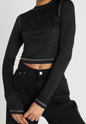 Frau trägt ein eng anliegendes, langärmliges schwarzes Crop-Top mit weißen Nähten und hoch taillierte schwarze Hose, die Hände vor dem Körper verschränkt.