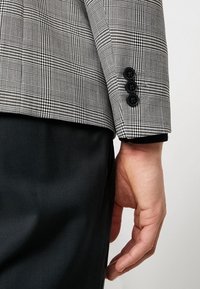 Grå rutigt kostymjacka ärm med tre svarta knappar, i kombination med ett par helsvarta byxor. Synlig textur och strukturerade design detaljer.