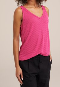 Kvinna i ett ärmlöst rosa strukturerat V-ringad topp, matchat med svarta byxor med dragsko, stående med en hand i fickan.