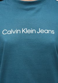 Roheline puuvillane T-särk, millel on ümar kaelus ja valge tekst "Calvin Klein Jeans" rinnal, siledast kangast.