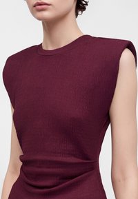 Personne portant un haut bordeaux texturé sans manches avec des épaules rembourrées et un col rond, présenté du cou à la taille.