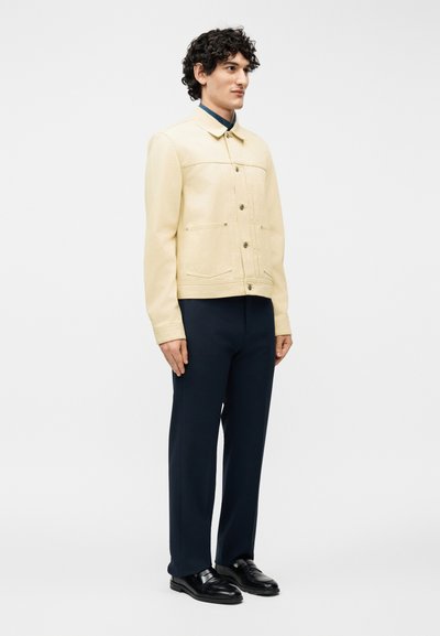 Άνδρας που στέκεται με το πρόσωπο προς τα αριστερά, φοράει μπεζ σακάκι με κουμπιά, σκούρο μπλε παντελόνι και μαύρα δερμάτινα μοκασίνια σε λευκό φόντο.