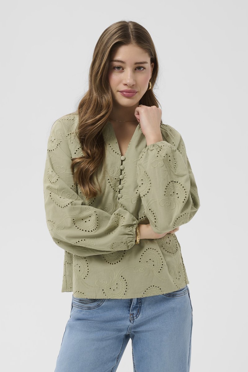 Ung kvinde med langt, bølget hår iført en lysegrøn broderet bluse med knapper og blå jeans, der poserer med hånden hvilende nær ansigtet.