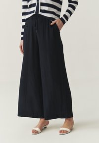 Pantalones de pierna ancha en color navy, fabricados con un material ligero y con una cinturilla ajustable. Combinados con sandalias blancas de punta abierta.