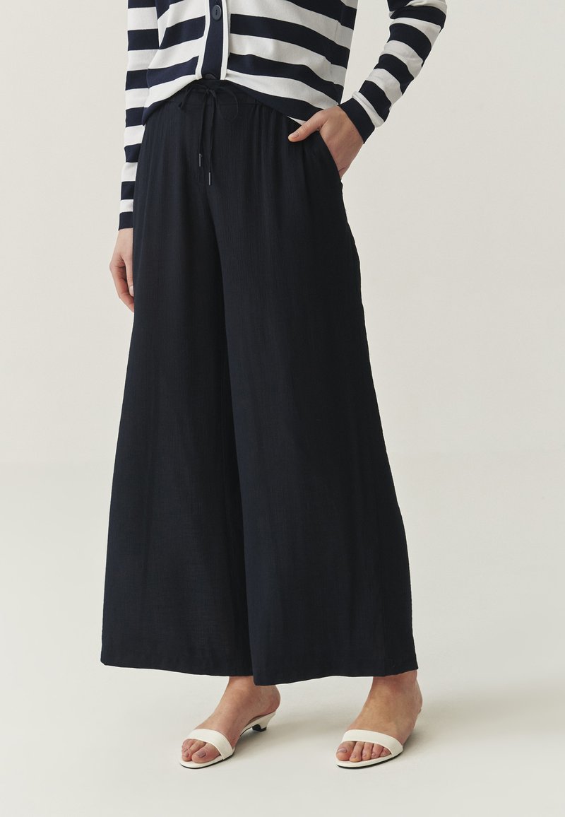 Pantalones de pierna ancha en color navy, fabricados con un material ligero y con una cinturilla ajustable. Combinados con sandalias blancas de punta abierta.