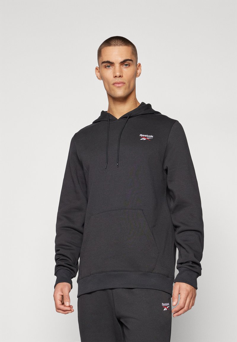 Haut à capuche noir en tissu doux, doté d'une poche frontale et d'une capuche avec cordon. Un petit logo Reebok rouge et blanc est présent sur la poitrine.