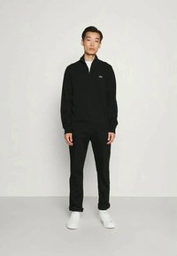 Pullover preto com fecho zíper, mangas longas, punhos canelados e um pequeno logótipo; combinado com calças pretas e ténis brancos. Design simples.