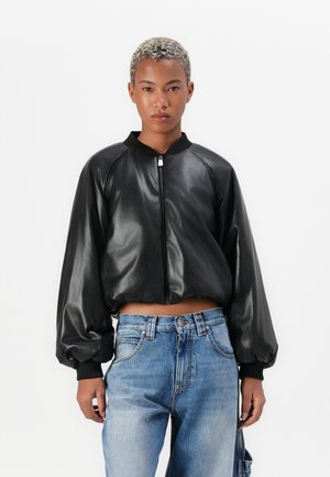 Giacca bomber nera cropped in pelle lucida con maniche a sbuffo e zip frontale. Abbinata a jeans in denimazzurro chiaro.