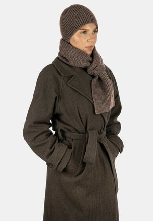 Frau trägt eine braune Strickmütze und einen passenden Schal, gekleidet in einem dunkelbraunen, mit Gürtel versehenen Wollmantel, die Hände in den Taschen, blickt zur Seite.