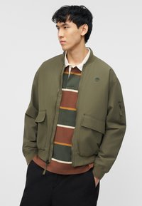 Νεαρός άνδρας που φοράει ένα χακί μπουφάν bomber πάνω από μια ριγωτή καφέ, πράσινη και χρυσή μπλούζα ράγκμπι, με μαύρο παντελόνι, τα χέρια στις τσέπες.