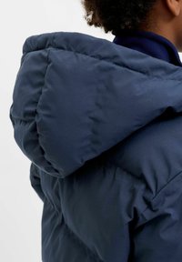 Tumši zila pūkaina jaka ar lielu kapuci, izceļot izšūtu audumu un gludu tekstūru. Apkakle ir augsta un kārtota, nodrošinot papildus siltumu.