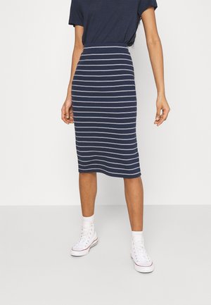 Kvinde iført en knælængde pencilskørt med marineblå og hvide striber, navy top og hvide høje sneakers, står mod en ensfarvet baggrund.