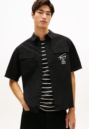 Młody mężczyzna ubrany w czarną koszulę na guziki z krótkim rękawem nałożoną na czarno-białą pasiastą koszulkę, stojący z jedną ręką w kieszeni.