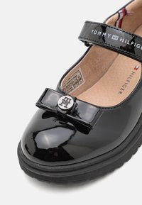 Zapato Mary Jane de charol negro con punta redonda, correa ajustable y un lazo decorativo que presenta un logo metálico en la parte frontal. Suela texturizada negra.