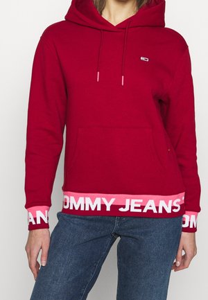 Sudadera con capucha roja fabricada en un tejido suave, que cuenta con un bolsillo frontal, cordones y una banda rosa en contraste con el texto "TOMMY JEANS".