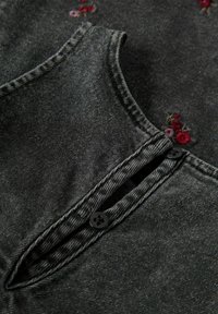 Svart denimtyg överdel med rundad krage, prydd med blommig brodyr och två svarta knappar nära halsringningen, som visar en texturerad yta.