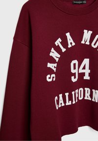Sweatshirt bordeaux avec texte blanc imprimé indiquant "SANTA MONICA 94 CALIFORNIA." Présente un col rond et un design ample et court.