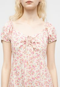 Ξανθιά γυναίκα που φοράει κρέμ φόρεμα με ροζ λουλουδάτο σχέδιο, φουσκωτά κοντά μανίκια και σούρες στο λαιμό με δέσιμο μπροστά.