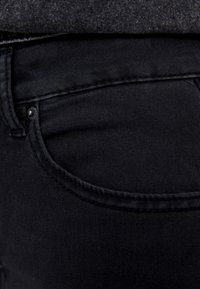 Vaqueros de denim negro con una textura suave, contorno de bolsillo curvado y detalle de botón de metal en la cintura. Diseño ajustado, sin adornos visibles.