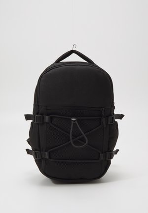 Sac à dos compact noir avec poche avant en filet, bretelles latérales réglables et poignée supérieure, debout sur un fond uni clair.