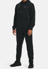 Zwarte sweatshirt- en broekset van zacht materiaal, met een voorzak, trekkoorden en paarse logo-accenten langs de benen.