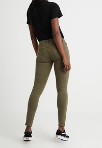 Persona vestida con jeans skinny verde oliva, camiseta negra y zapatillas Vans negras de pie, de espaldas a la cámara.