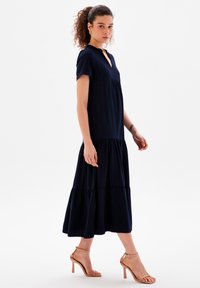 Vestido midi azul marino hecho de tela ligera, con mangas cortas, escote en V y volantes en capas en el dobladillo. Combinado con sandalias nude de tiras.