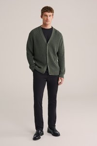 Zelený žebrovaný pletený cardigan s hlubokým výstřihem do V a zapínáním na knoflíky, nošený přes černou košili, doplněný černými kalhotami a černými botami.