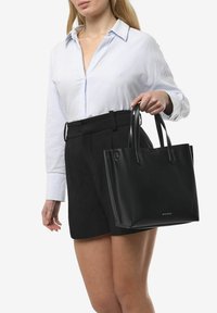Zwarte leren tote bag met een gladde textuur, rechthoekige vorm en dubbele handgrepen. Heeft een minimalistisch ontwerp en subtiele branding.