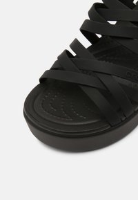 Sandale en caoutchouc noir avec des lanières entrecroisées, une semelle intérieure texturée et une semelle rembourrée. Design à bout fermé avec une légère élévation au talon.