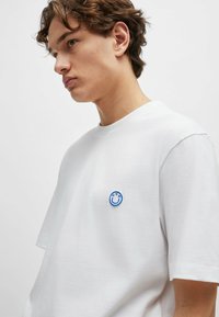 White cotton t-shirt with short sleeves, featuring a blue embroidered smiley face logo on the left chest. Smooth texture and loose fit.