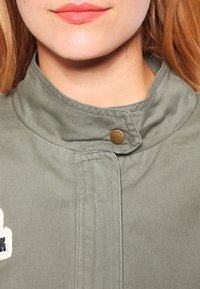 Primer plano de la parte inferior de la cara y el cuello de una mujer que lleva una chaqueta verde de cuello alto, con un botón de metal abrochado cerca del mentón.