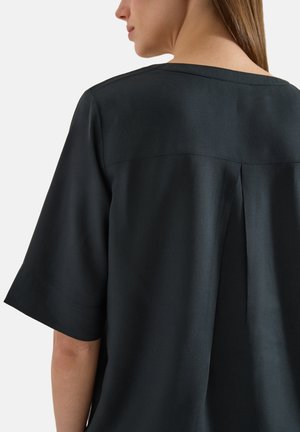 Femme portant un haut noir ample avec un pli creux dans le dos et des manches aux coudes, vue de dos.