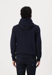 Uomo con capelli ricci che indossa un hoodie blu navy e jeans scuri, in piedi di spalle contro uno sfondo bianco semplice.