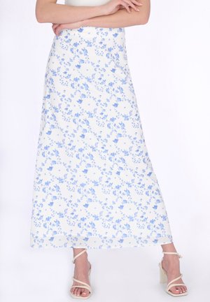 Vrouw draagt een witte midi-rok met een kleine blauwe bloemenprint, gecombineerd met witte sandalen met hoge hak en bandjes, armen over elkaar boven de taille.