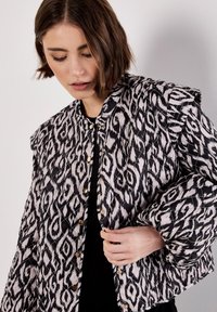 Quiltad jacka i svart och vitt med ett abstrakt mönster, med rund halsringning och guldknappar fram.