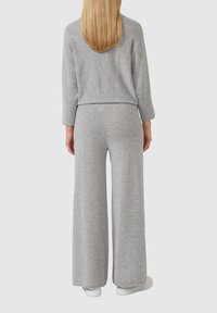 Femme aux longs cheveux blonds portant un haut gris à manches longues et un pantalon large, se tenant de dos avec des baskets blanches.