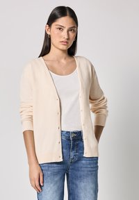 Cardigan color beige chiaro con scollo a V, maniche lunghe e polsini a coste. Presenta bottoni frontali ed è indossato su una canotta bianca con jeans blu.